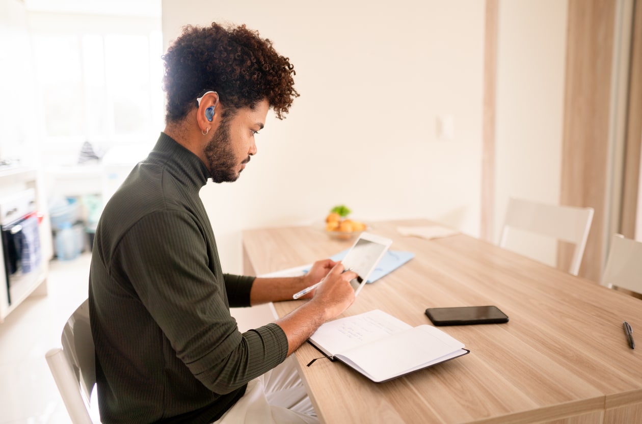 Young man with a hearing aid working on a tablet at home Man connecting hearing aid to his tablet.