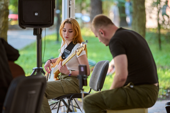 yung woman plays guitar while man drums outdoors. Trees and warm lighting create relaxed musical atmosphere
